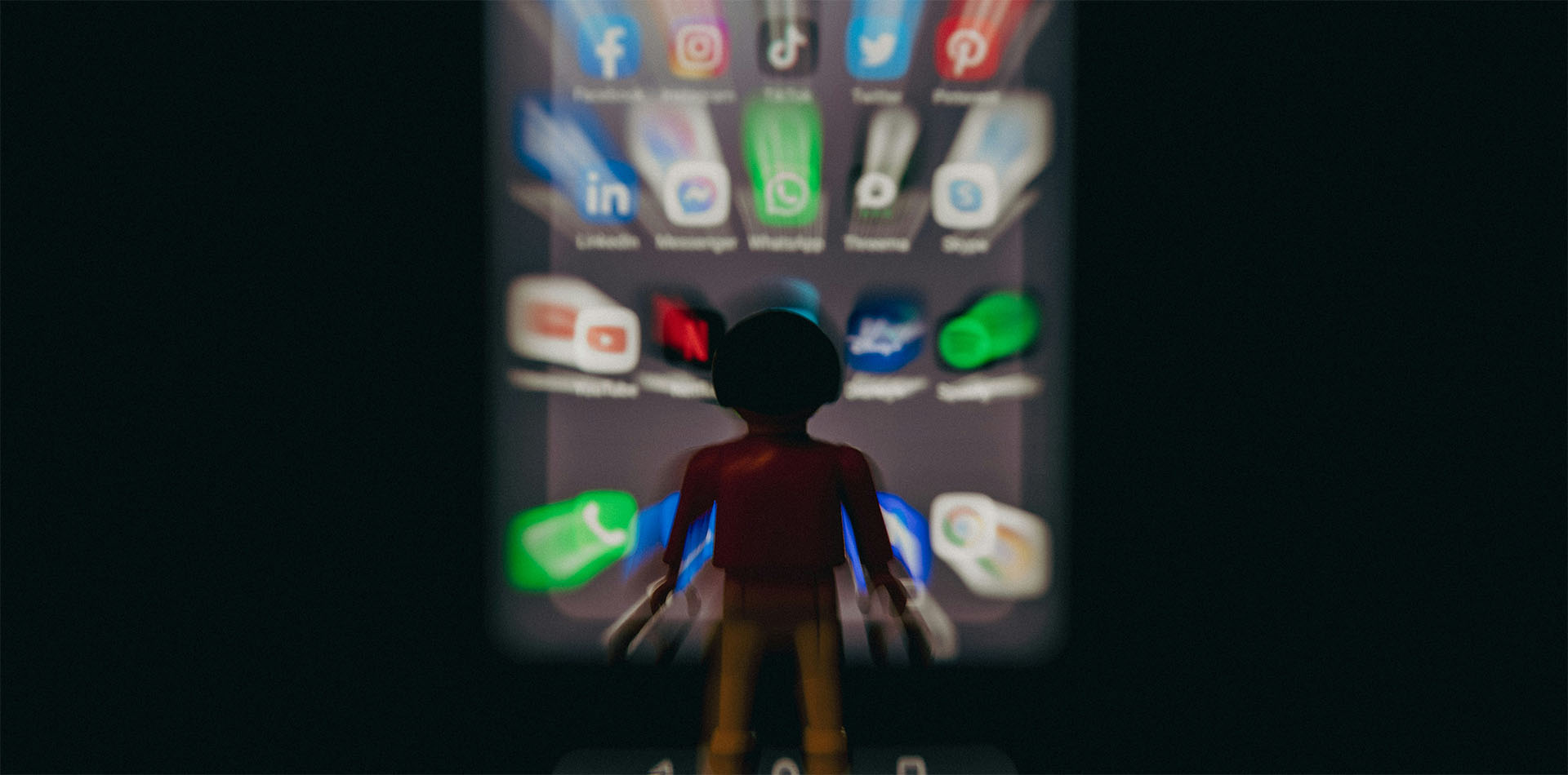 Toy figure facing a glowing smartphone screen filled with blurred social media app icons.